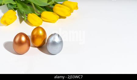 Trois œufs de Pâques peints de plusieurs couleurs sur une table à côté de tulipes jaunes sur fond blanc. Vacances de printemps. Vacances de Pâques. Minimalisme Banque D'Images