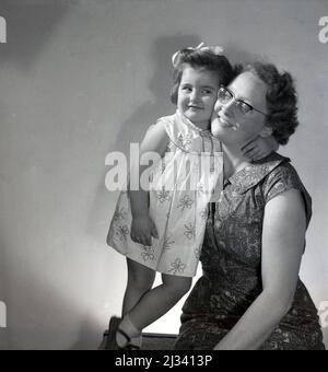 1961, historique, mère et enfant, une petite fille dans une robe et un arc dans ses cheveux, donnant à sa mère une câlin aimante, Stockport, Manchester, Angleterre, Royaume-Uni. Banque D'Images