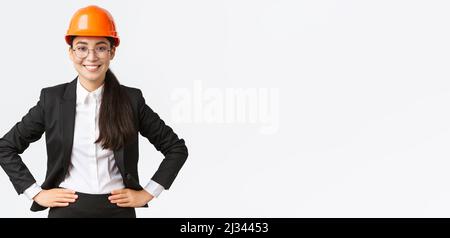 Femme asiatique sourit, ingénieur de construction, architecte en costume noir et casque de sécurité, qui se tient satisfaite de son travail Banque D'Images