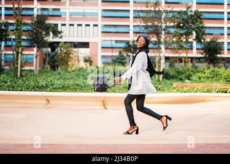 Vue latérale sur tout le corps de la jeune femme afro-américaine élégante dans un sac balançoire traditionnel avec foulard tout en se promenant sans ménagé dans un parc urbain et en regardant loin Banque D'Images