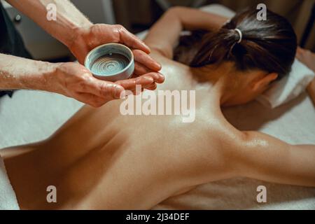 Un bol avec de l'huile chaude infusée aux herbes pour le massage du dos dans les mains d'un thérapeute masculin Banque D'Images