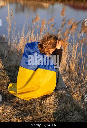 Pleurant jeune femme patriote malheureuse. Enveloppée dans le drapeau ukrainien se trouve sur le bord du lac dans le désespoir. Guerre en Ukraine, agression russe. Souffrent Banque D'Images