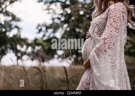 Femme enceinte aux cheveux longs blonds dans la forêt, portant une robe en dentelle boho. Belle robe et s'attendant à une femme à l'extérieur dans la nature. Banque D'Images