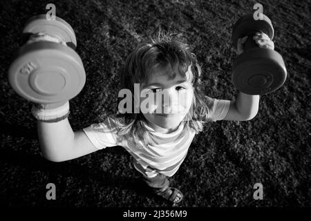 Sport amusant pour enfants. Enfant avec haltères dans le parc. Petit garçon fort. Visage d'enfant amusant. Développement des enfants et exercice sain et fort. Grand angle. Banque D'Images