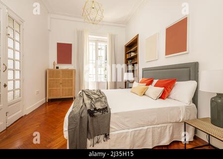 Chambre avec lit double avec coussins orange, tête de lit rembourrée en tissu gris avec clous dorés, buffets en osier et balcon avec doo en bois blanc Banque D'Images