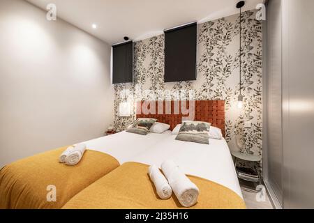 Chambre avec lit double avec tête de lit en cuir tressé, mur recouvert de papier peint décoratif, lampes de suspension, couvertures de couleur moutarde et placard Banque D'Images