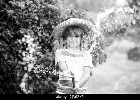 Un joli petit garçon en chapeau de paille rit avec un tuyau de pulvérisation d'eau. Banque D'Images