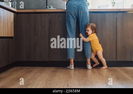 Portrait d'une petite fille embrassant la jambe de la mère et demandant à prendre. Pieds de maintien pour bébé debout. Soins familiaux. Soins de bébé. Banque D'Images