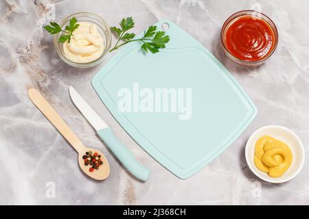 Bols en porcelaine et en verre avec ketchup, moutarde et sauce au fromage avec des feuilles de persil et un couteau, une cuillère en bois avec des épices sur la table de cuisine. Banque D'Images