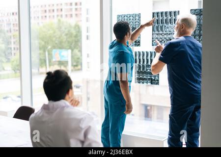 Vue arrière des médecins multi-ethniques de l'équipe discutant ensemble de l'histoire de la maladie du patient à l'aide de l'IRM pendant la conférence médicale debout près de la fenêtre. Banque D'Images