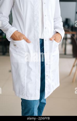Gros plan vertical de femme médecin méconnaissable en uniforme blanc tenant les mains dans les poches debout dans le bureau de l'hôpital. Banque D'Images