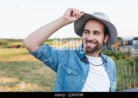 jeune et heureux fermier touchant chapeau de brime tout en regardant loin dans le champ Banque D'Images