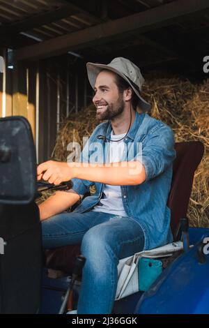 joyeux fermier en chapeau de brime regardant loin en étant assis sur le tracteur Banque D'Images