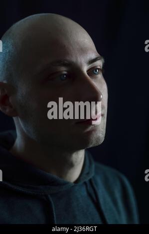 Portrait d'un beau jeune homme chauve aux yeux bleus qui, dans la faible lumière, regarde soigneusement loin Banque D'Images