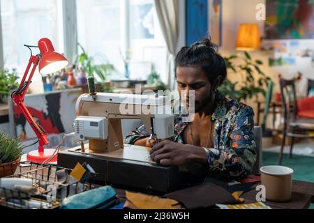 Portrait du jeune Indien créatif assis à une table dans le salon à la maison en utilisant la machine à coudre pour faire des vêtements neufs Banque D'Images