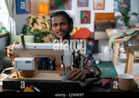 Portrait du jeune Indien joyeux avec le nez pierant assis devant la machine à coudre électrique faisant de nouveaux vêtements collection Banque D'Images