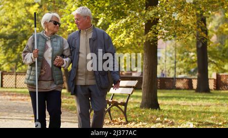 La femme aveugle caucasienne senior avec des lunettes et un bâton s'appuie sur son mari tout en ayant une promenade dans le parc handicapés aveugles personnes prennent en charge concept moyen plein espace de copie de tir. Photo de haute qualité Banque D'Images