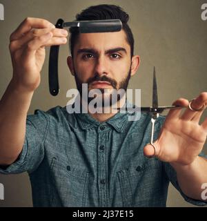 Tondez Oui rasage non. Photo en studio d'un jeune homme charmant tenant un rasoir droit et un ciseaux entre ses mains. Banque D'Images