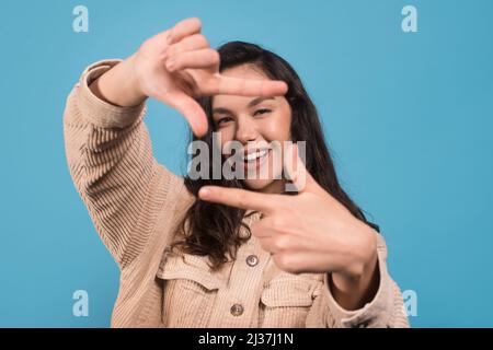 Jeune gaie jolie femme européenne regardant à travers les doigts comme un appareil photo isolé sur fond bleu Banque D'Images