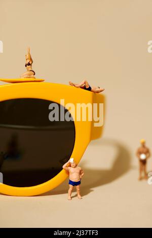 quelques personnes miniatures, portant un maillot de bain, autour et sur une paire de lunettes de soleil jaunes à rebord en plastique sur un fond beige Banque D'Images