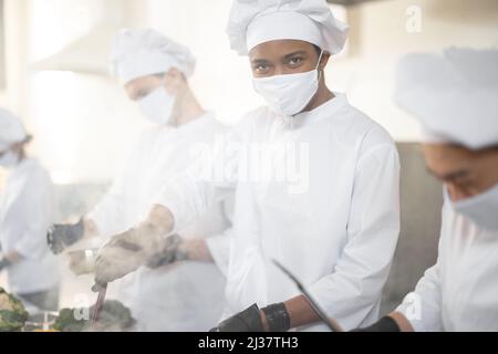 Une équipe multiraciale de cuisiniers cuisiniers en uniforme blanc dans la cuisine. Chef asiatique avec cuisine latine friture de viande, les gars européens cuisinent sur fond. Cuisiniers portant des masques et des gants de protection Banque D'Images