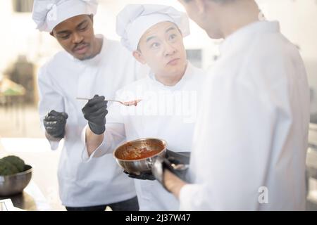 Trois cuisiniers cuisiniers avec différentes origines sauce dégustation avec une cuillère tout en cuisant dans la cuisine. Travail d'équipe et cuisine délicieuse au restaurant Banque D'Images