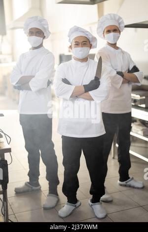Portrait de trois cuisiniers de chef bien habillés avec différentes origines ethniques debout ensemble dans la cuisine du restaurant. Chef asiatique tenant le couteau, latins et européens sur fond. Les cuisiniers portant des masques faciaux prêts à cuire pendant une pandémie Banque D'Images