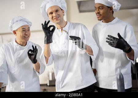 Trois cuisiniers avec différentes spécialités goûtant des plats cuisinés, inventant de nouveaux plats pour un menu. Chef caucasien avec des cuisiniers latins et asiatiques en uniforme travaillant ensemble. Concept de travail d'équipe et cuisine haute Banque D'Images