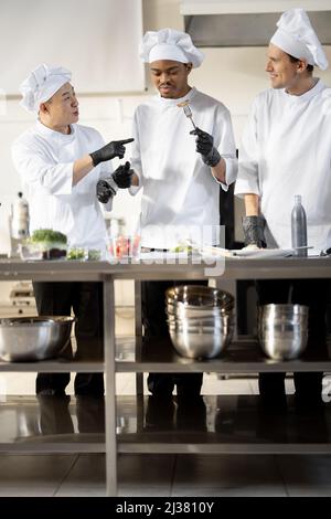 Trois cuisiniers avec différentes spécialités goûtant des plats cuisinés, inventant de nouveaux plats pour un menu. Chef asiatique avec des cuisiniers latino et européens en uniforme travaillant ensemble. Concept de travail d'équipe et cuisine haute Banque D'Images