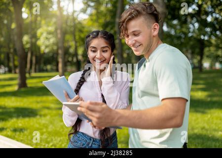 Les étudiants multiraciaux débordient de joie en vérifiant les résultats des examens sur une tablette numérique et en souriant, en marchant dans un parc ou sur un campus universitaire Banque D'Images