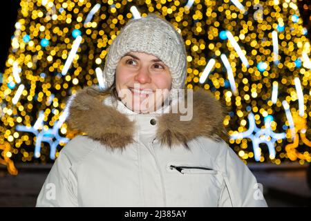 Une femme heureuse dans un chapeau blanc et une veste se tient près d'un arbre de Noël, bokeh jaune, à l'extérieur Banque D'Images