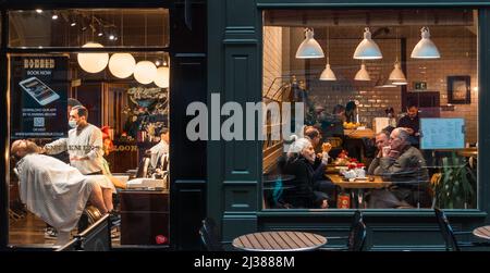 Deux vitrines contrastées très fréquentées dans le centre-ville de Leeds - un magasin de coiffure avec un homme se rasent et des gens qui boivent du café dans un café, West Yorkshire, Angleterre, Royaume-Uni Banque D'Images