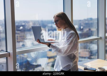 Femme d'affaires élégante en lunettes pour la correction de la vue, elle tient un ordinateur portable moderne dans ses mains, debout près d'une fenêtre pleine-mur dans un moderne Banque D'Images