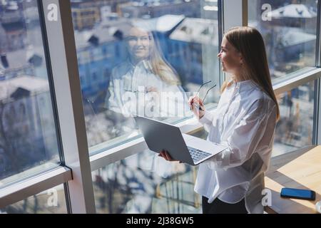 Femme d'affaires élégante en lunettes pour la correction de la vue, elle tient un ordinateur portable moderne dans ses mains, debout près d'une fenêtre pleine-mur dans un moderne Banque D'Images
