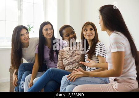 Groupe diversifié de jeunes femmes heureux assis sur un canapé à la maison, parlant et partageant des nouvelles Banque D'Images
