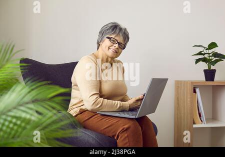 Portrait d'une femme âgée heureuse assise dans un fauteuil à la maison avec son ordinateur portable Banque D'Images