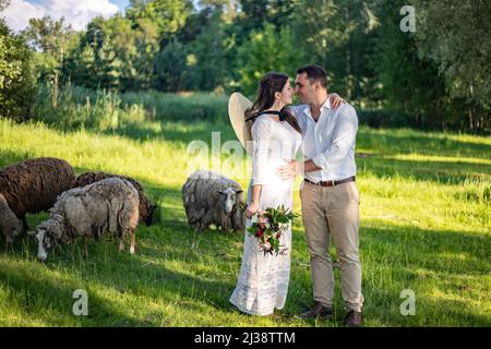 De magnifiques jeunes mariés se tiennent sur le fond d'une clôture à feuillage vert. L'élégant marié tient la main de la mariée. Mariage Banque D'Images