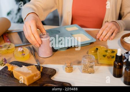 Vue rognée de l'artisan prenant pot avec du savon près de moule en silicone, les huiles essentielles et les fleurs sur la table Banque D'Images