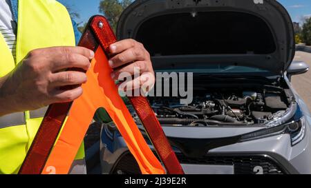Voiture avec des problèmes et un triangle rouge pour avertir les autres usagers de la route. Conducteur mâle se tenant près d'une voiture brisée avec capot ouvert. Banque D'Images