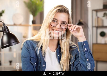 Femme caucasienne souriante en lunettes et chemise en denim, posant à l'intérieur à l'intérieur lumineux de bureau ou à l'intérieur de la maison. Jolie dame ajuster ses lunettes et sourire sur l'appareil photo Banque D'Images