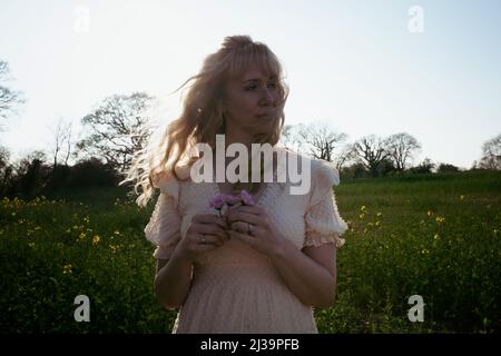 femme blonde aux cheveux courbés tenant des fleurs dans un champ au coucher du soleil Banque D'Images