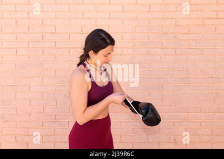 Une jeune femme caucasienne vêtue d'un haut met un gant de boxe noir devant un mur de briques. Banque D'Images