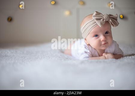 Bébé fille de 3 mois se trouve sur un lit avec des vêtements blancs sur son ventre et sourit, regarde la caméra, bébé matin, le concept de substance de bébé. Banque D'Images