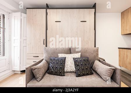 canapé-lit en tissu gris avec coussins bleus et blancs, balcon avec volets en bois et armoire intégrée dans le studio de location de vacances Banque D'Images