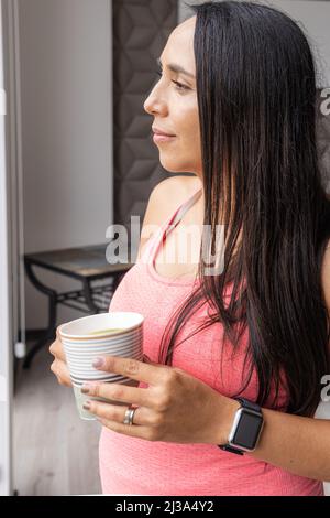 Plan vertical d'une jeune femme enceinte qui boit une tasse d'eau aromatique en regardant par la fenêtre Banque D'Images