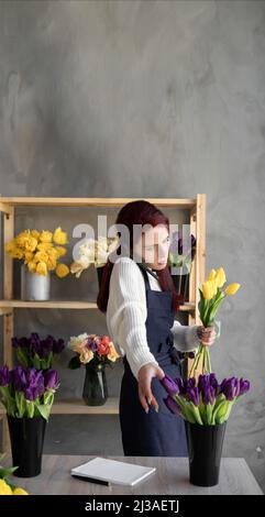 Concept de petite entreprise. Une jeune belle femme fleuriste dans un tablier se tient près d'une vitrine avec des tulipes prend une commande par téléphone et recueille un bouquet. Banque D'Images