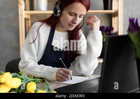 Jeune belle fleuriste femelle dans le tablier bleu propriétaire d'affaires en utilisant le casque et l'ordinateur tout en parlant à des clients en ligne prenant la commande en ligne. Banque D'Images