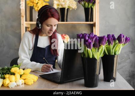 Jeune femme fleuriste en tablier bleu propriétaire d'affaires utilisant le casque et l'ordinateur tout en parlant avec les clients en ligne prend commande en ligne, écrire avec Banque D'Images