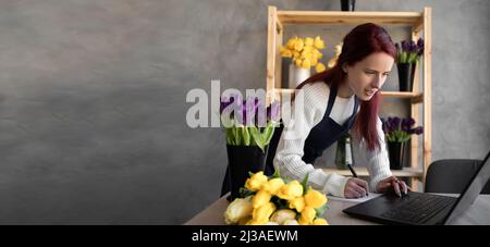 Portrait d'une jeune fille fleuriste dans un tablier bleu regardant les commandes des clients sur un ordinateur portable. Concept pour la livraison de fleurs et la logistique de petite entreprise. Banque D'Images