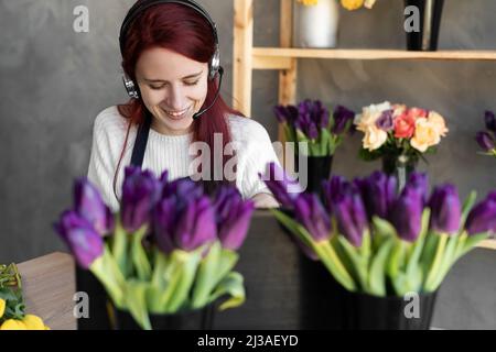 Jeune femme fleuriste en tablier bleu propriétaire d'affaires utilisant le casque et l'ordinateur tout en parlant avec les clients en ligne prend commande en ligne, écrire avec Banque D'Images